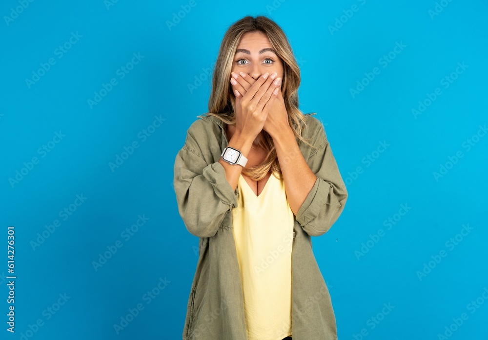Stunned young beautiful blonde woman standing over blue studio background covers both hands on mouth, afraids of something astonishing Stunned young beautiful blonde woman standing over blue studio background covers both hands on mouth, afraids of something astonishing