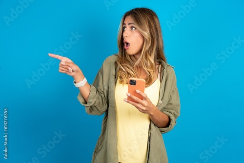 Stunned young beautiful blonde woman standing over blue studio background points sideways right copy space, recommends product, sees astonishing thing photo