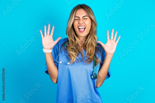 Emotive Young beautiful doctor woman standing over blue studio background laughs loudly, hears funny joke or story, raises palms with satisfaction,