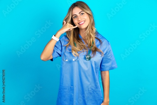 Young beautiful doctor woman standing over blue studio background smiling doing phone gesture with hand and fingers like talking on the telephone. Communicating concepts.