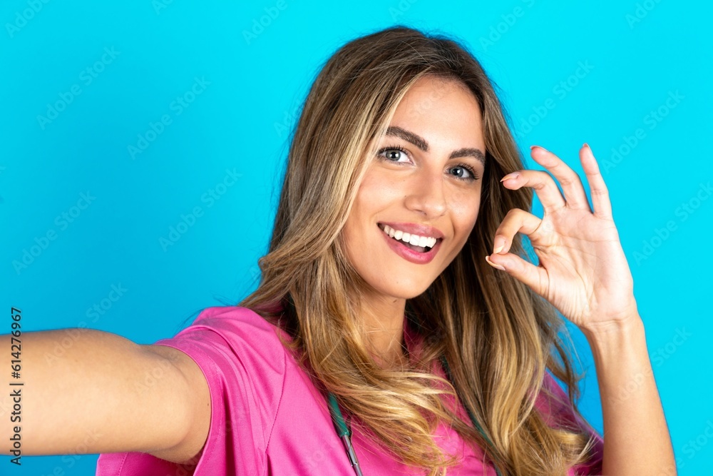 Portrait of pretty cheerful Young beautiful doctor woman standing over blue studio background make selfie show okey symbol Portrait of pretty cheerful Young beautiful doctor woman standing over blue studio background make selfie show okey symbol