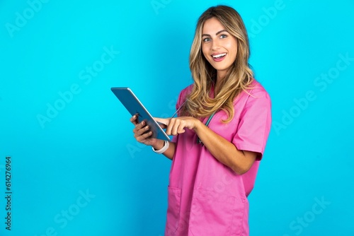Photo of happy cheerful smart Young beautiful doctor woman standing over blue studio background hold tablet browsing internet