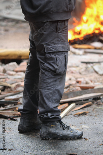 detalhe de pernas de homem vestindo calca com bolsos laterais e botas de trabalho em escombros de explosao e incendio ao fundo. cena de filmagens de atentado.