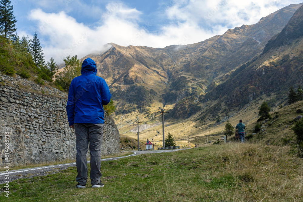 A tourist in a blue jacket looks at the road in the mountains, back view. Carpathians, Ridge Fagarash. Transfagarash road
