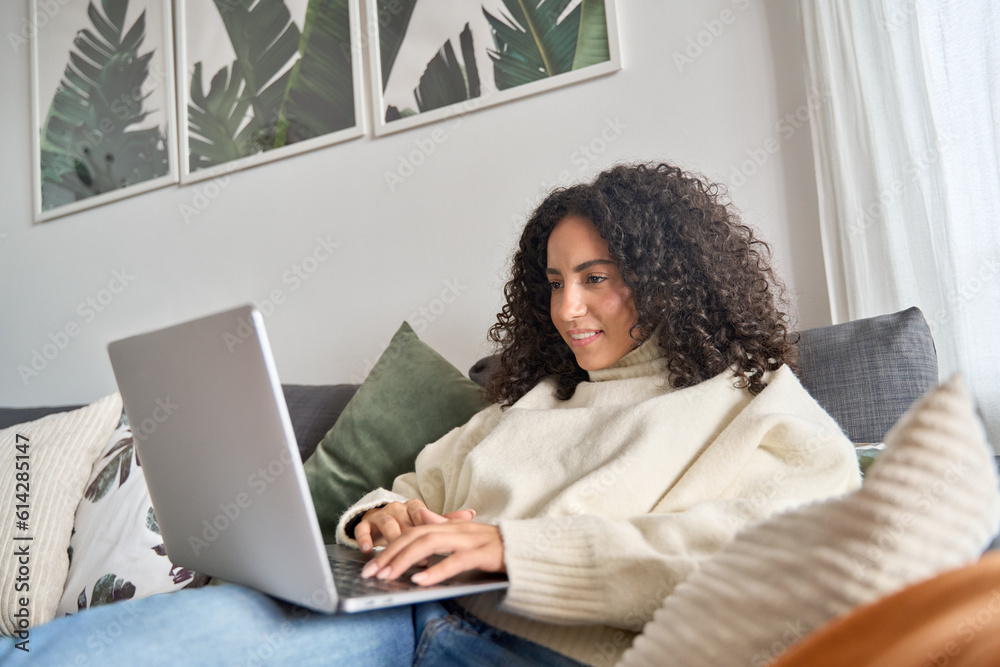 Relaxed latin young woman sitting on sofa using laptop at home surfing, doing online ecommerce shopping, looking at computer buying sale products on website, watching videos or elearning. Relaxed latin young woman sitting on sofa using laptop at home surfing, doing online ecommerce shopping, looking at computer buying sale products on website, watching videos or elearning.
