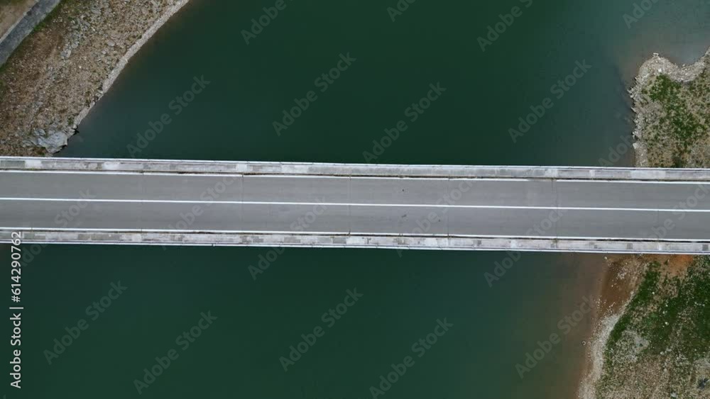 Aerial view of a bridge over a river, the road horizontally separates the image in half, a grey car crosses the frame from left to right, the drone moves down zooming in slightly.