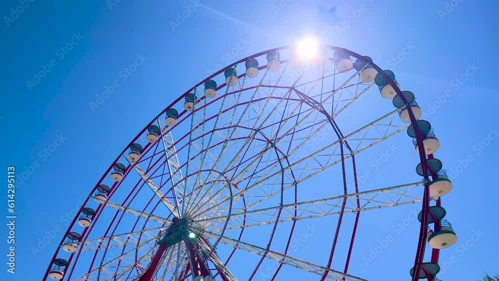 Wide angle shot: view of palm trees and ferris wheel is rotating against clear blue sky with sun lens flare at amusement park. Sightseeing, cityscape, entertainment and holiday concept Wide angle shot: view of palm trees and ferris wheel is rotating against clear blue sky with sun lens flare at amusement park. Sightseeing, cityscape, entertainment and holiday concept