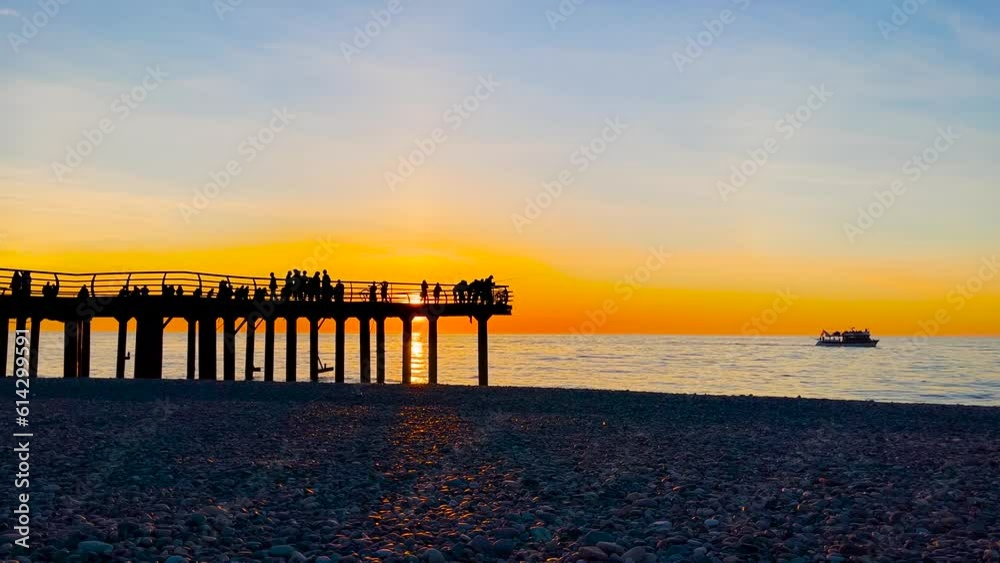 Unrecognizable silhouettes of people are walking on the pier on the pebble beach against the dramatic sky over the Black Sea at sunset: slow motion, wide shot. Summer, vacation and tropical concept
