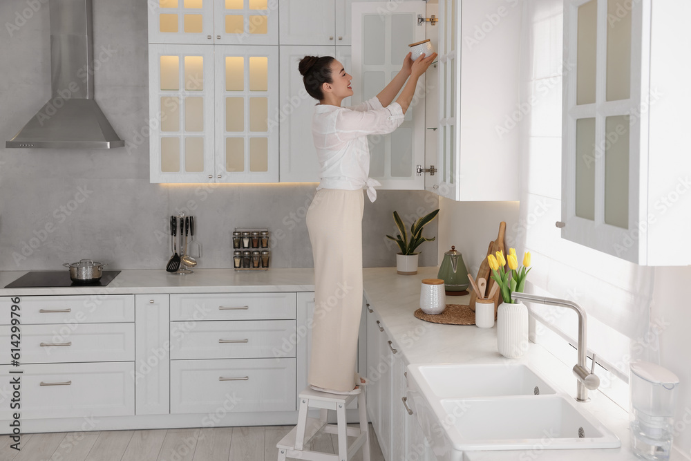 Woman on ladder putting white jar into cupboard at home Woman on ladder putting white jar into cupboard at home