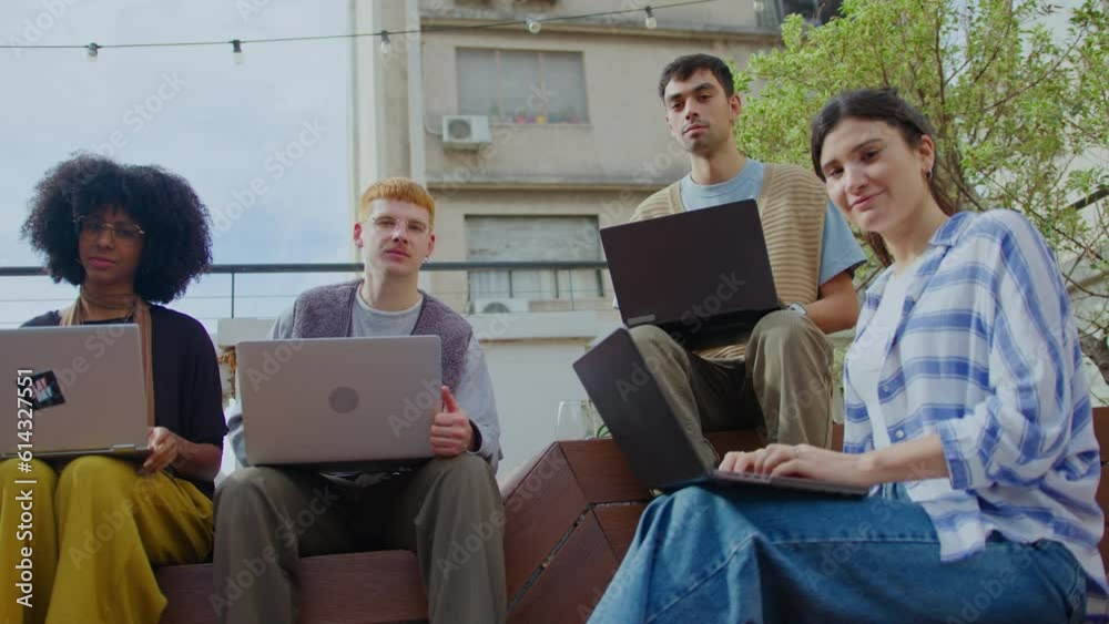 Team of diverse gen Z business people in casual wear sitting with laptops on bench in the city, posing together on camera during the day. Zoom shot, group portrait Team of diverse gen Z business people in casual wear sitting with laptops on bench in the city, posing together on camera during the day. Zoom shot, group portrait