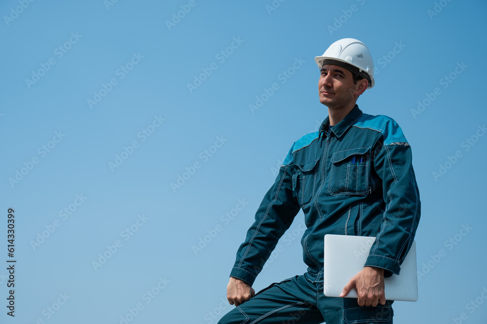 A male builder in a helmet stands against a blue sky with a laptop in his hands. A male builder in a helmet stands against a blue sky with a laptop in his hands.