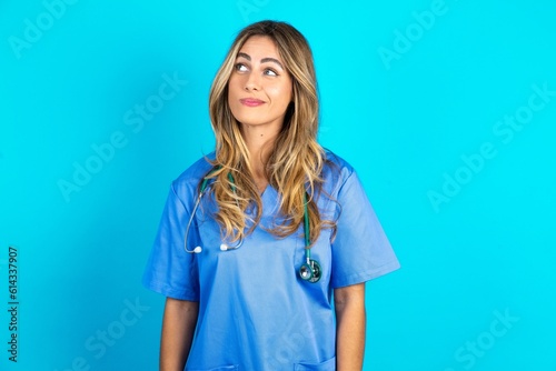 Young caucasian blonde doctor woman wearing blue uniform looking aside into empty space thoughtful