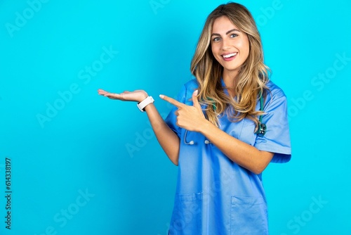 Positive Young caucasian blonde doctor woman wearing blue uniform promoter point index finger copyspace hold hand demonstrate offer ads promo