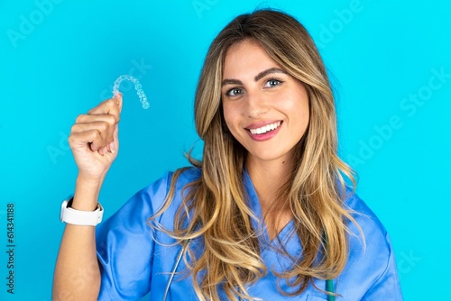 Young caucasian blonde doctor woman wearing blue uniform holding an invisible braces aligner, recommending this new treatment. Dental healthcare concept.