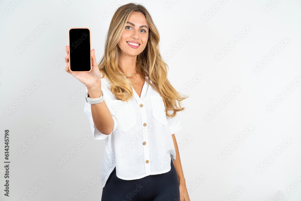 Smiling young blonde woman wearing white shirt over white studio background Mock up copy space. Hold mobile phone with blank empty screen