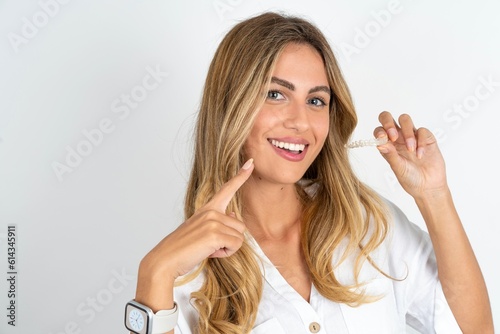 young blonde woman wearing white shirt over white studio background holding an invisible aligner and pointing to her perfect straight teeth. Dental healthcare and confidence concept.