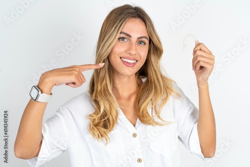 young blonde woman wearing white shirt over white studio background holding an invisible aligner and pointing perfect straight teeth. Dental healthcare and confidence concept.