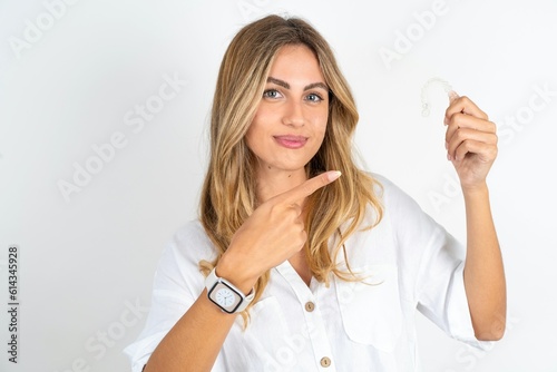 young blonde woman wearing white shirt over white studio background holding an invisible aligner and pointing at it. Dental healthcare and confidence concept.