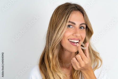 young blonde woman wearing white shirt over white studio background holding an invisible aligner ready to use it. Dental healthcare and confidence concept.