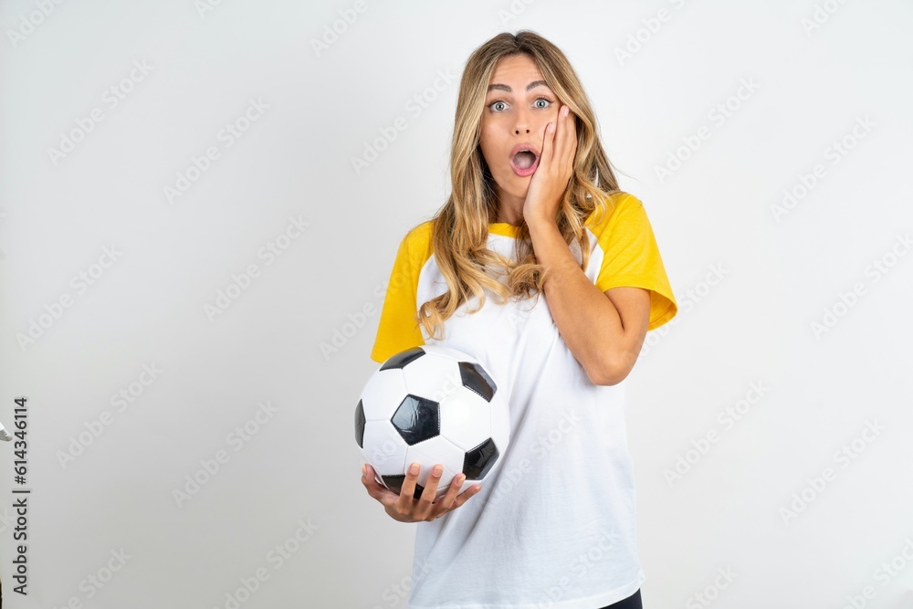 Speechless Young beautiful woman wearing football T-shirt over white background keeps hands near opened mouth reacts to shocking news stares wondered at camera Speechless Young beautiful woman wearing football T-shirt over white background keeps hands near opened mouth reacts to shocking news stares wondered at camera