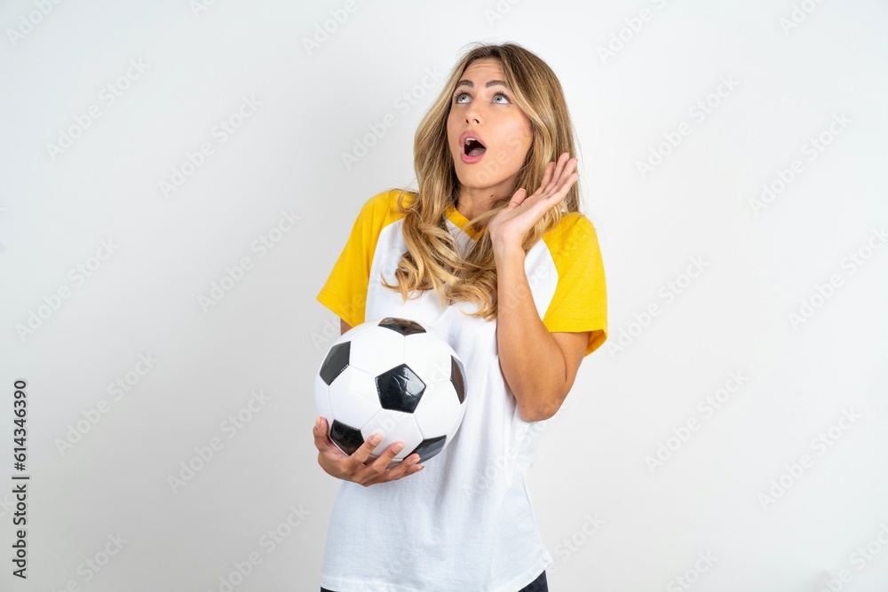Young beautiful woman wearing football T-shirt over white background keeps palms forward and looks with fright above on ceiling tries to defense herself from invisible danger opens mouth. Young beautiful woman wearing football T-shirt over white background keeps palms forward and looks with fright above on ceiling tries to defense herself from invisible danger opens mouth.