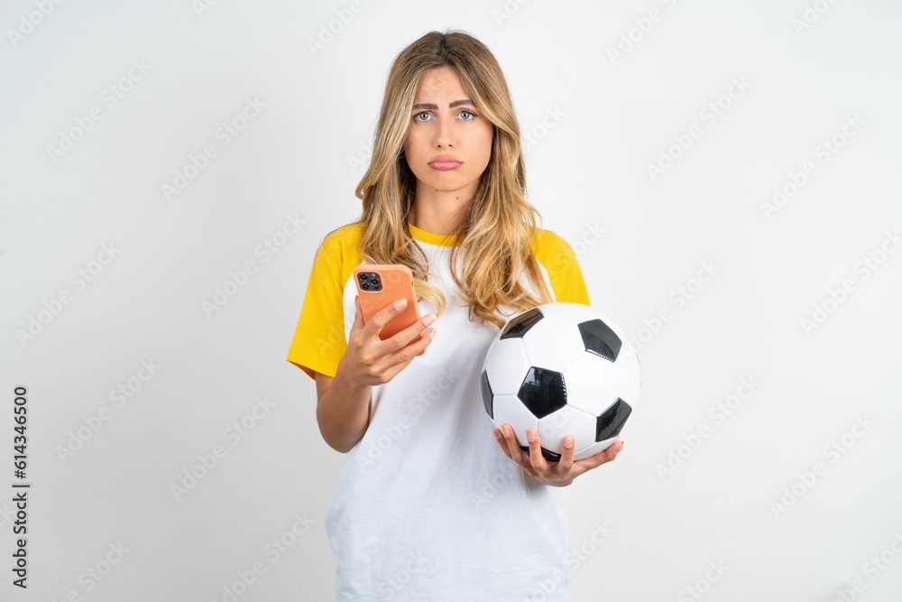 Upset dissatisfied Young beautiful woman wearing football T-shirt over white background uses mobile software application and surfs information in internet, holds modern mobile hand Upset dissatisfied Young beautiful woman wearing football T-shirt over white background uses mobile software application and surfs information in internet, holds modern mobile hand