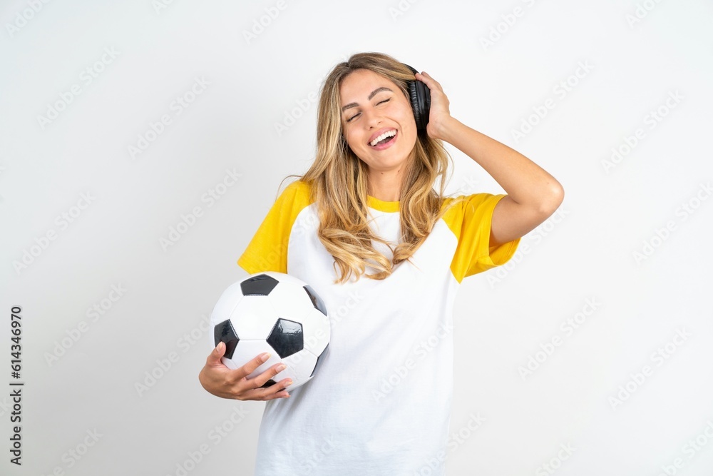 Young beautiful woman wearing football T-shirt over white background smiles broadly feels very glad listens favourite music track via wireless headphones closes eyes. Young beautiful woman wearing football T-shirt over white background smiles broadly feels very glad listens favourite music track via wireless headphones closes eyes.