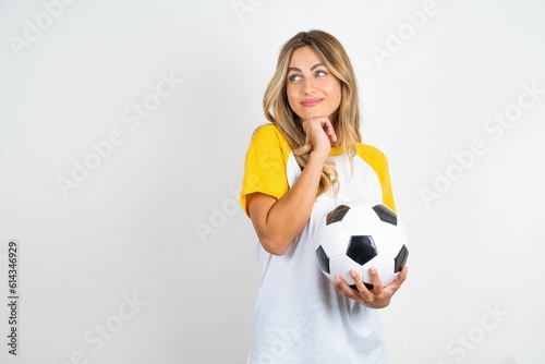 Curious Young beautiful woman wearing football T-shirt over white background keeps hands under chin bites lips and looks with interest aside.