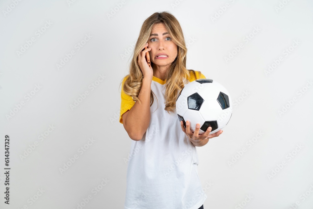 Afraid funny Young beautiful woman wearing football T-shirt over white background holding telephone and bitting nails Afraid funny Young beautiful woman wearing football T-shirt over white background holding telephone and bitting nails