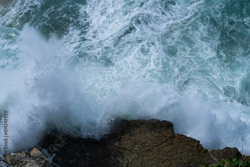 Big waves crashing into black rocks, blue sky background in Colares, Portugal. Ocean waves crashing against cliffs. Top down seashore. Waves crashing on rock cliff. Beautiful dark sea in Portugal.