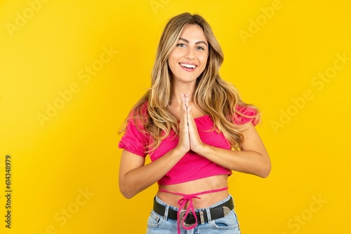 young blonde woman wearing pink crop top over yellow studio background praying with hands together asking for forgiveness smiling confident. photo
