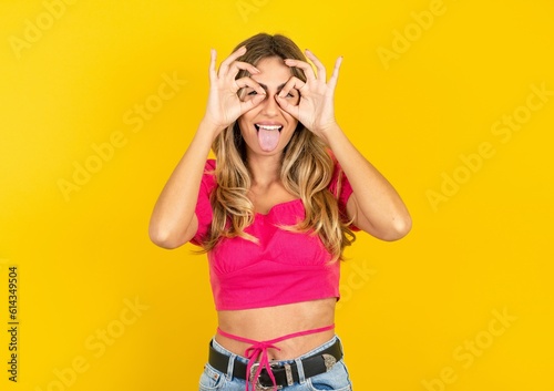 young blonde woman wearing pink crop top over yellow studio background doing ok gesture like binoculars sticking tongue out, eyes looking through fingers. Crazy expression.