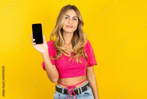 Smiling young blonde woman wearing pink crop top over yellow studio background Mock up copy space. Hold mobile phone with blank empty screen