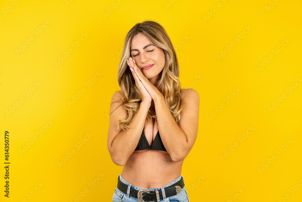 Overjoyed young beautiful blonde woman wearing bikini over yellow studio background laughs joyfully and keeps palms pressed together hears something funny Overjoyed young beautiful blonde woman wearing bikini over yellow studio background laughs joyfully and keeps palms pressed together hears something funny