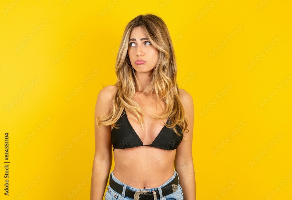 Dissatisfied young beautiful blonde woman wearing bikini over yellow studio background purses lips and has unhappy expression looks away stands offended. Depressed frustrated model.