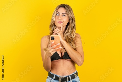 young beautiful blonde woman wearing bikini over yellow studio background thinks deeply about something, uses modern mobile phone, tries to made up good message, keeps index finger near lips.