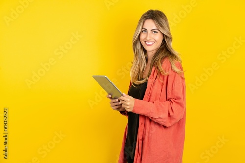 Photo of optimistic young beautiful blonde woman standing over yellow studio background hold tablet