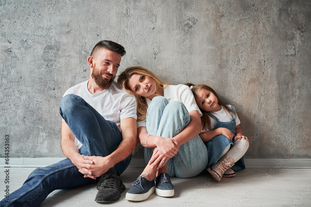 Adorable little girl and parents sitting by the wall in new apartment. Woman leaning head on husband shoulder while resting with family in new home.