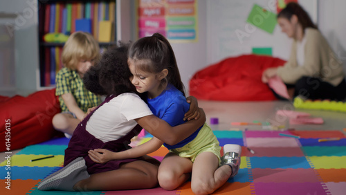 Fotografie Cute diverse little girls hug sitting on carpet in kindergarten