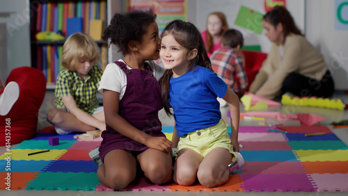 Multiracial little girls tell secrets sitting on floor in kindergarten