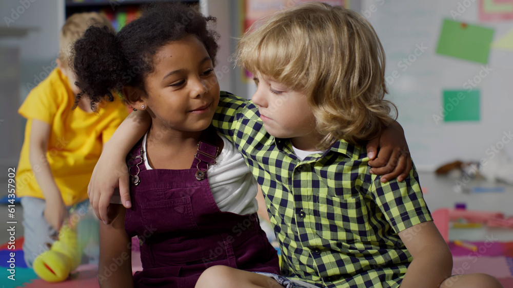 Adorable diverse kids talk and hug sitting on floor in kindergarten playroom Adorable diverse kids talk and hug sitting on floor in kindergarten playroom