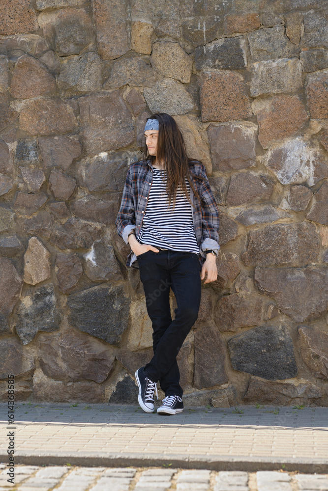 Young long-haired man in plaid shirt, striped t-shirt and jeans posing against stone wall Young long-haired man in plaid shirt, striped t-shirt and jeans posing against stone wall