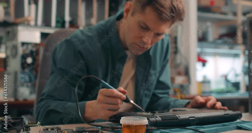 young man working with broken disassembled laptop. Electronic components. Computer analysis. Slow motion attractive man using a soldering iron to fix broken hardware at the repair shop. young man working with broken disassembled laptop. Electronic components. Computer analysis. Slow motion attractive man using a soldering iron to fix broken hardware at the repair shop.