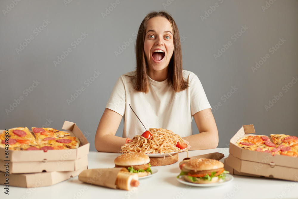 Amazed excited woman with brown hair wearing white T-shirt sitting at table among unhealthy food isolated over gray background breaks diet looking at camera screaming with cheerful face.