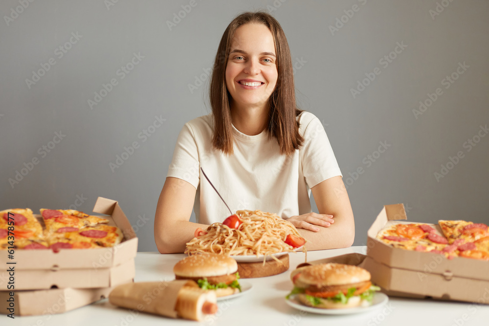 Smiling joyful young caucasian woman with brown hair wearing white T-shirt sitting at table among fast food isolated over gray background likes to eat unhealthy dishes.