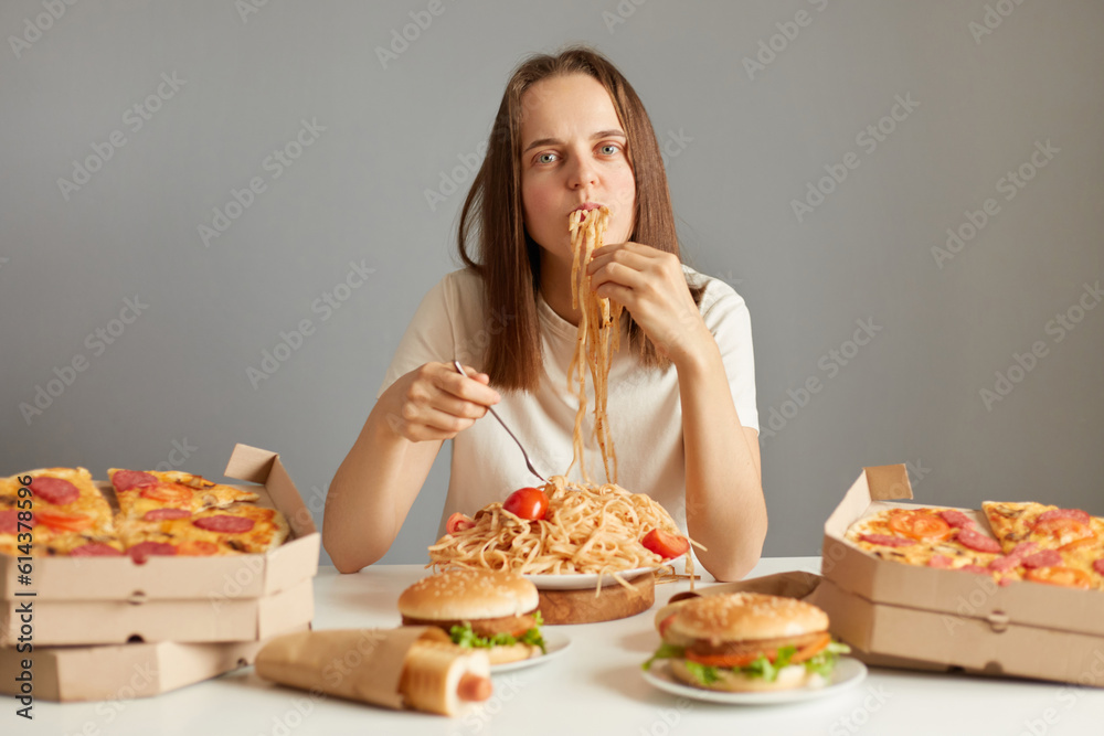 Funny woman with brown hair wearing white T-shirt sitting at table among unhealthy food isolated over gray background sitting with mouth full of pasta looking at camera.