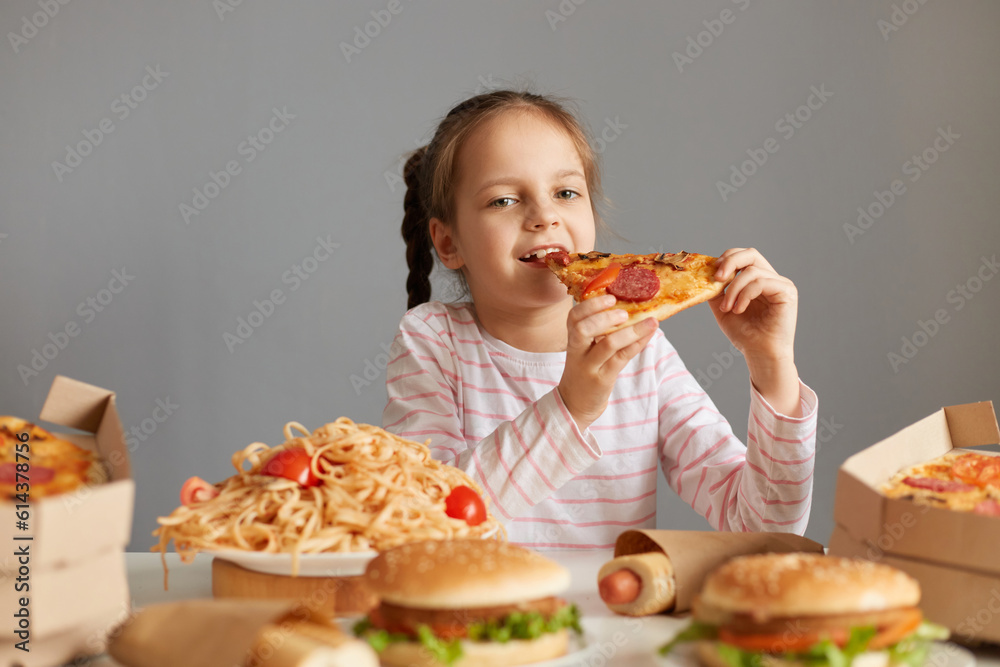 Hungry optimistic attractive little girl with braids sitting at table with junk food isolated over gray background biting big slice of pizza enjoying good appetite dinner.