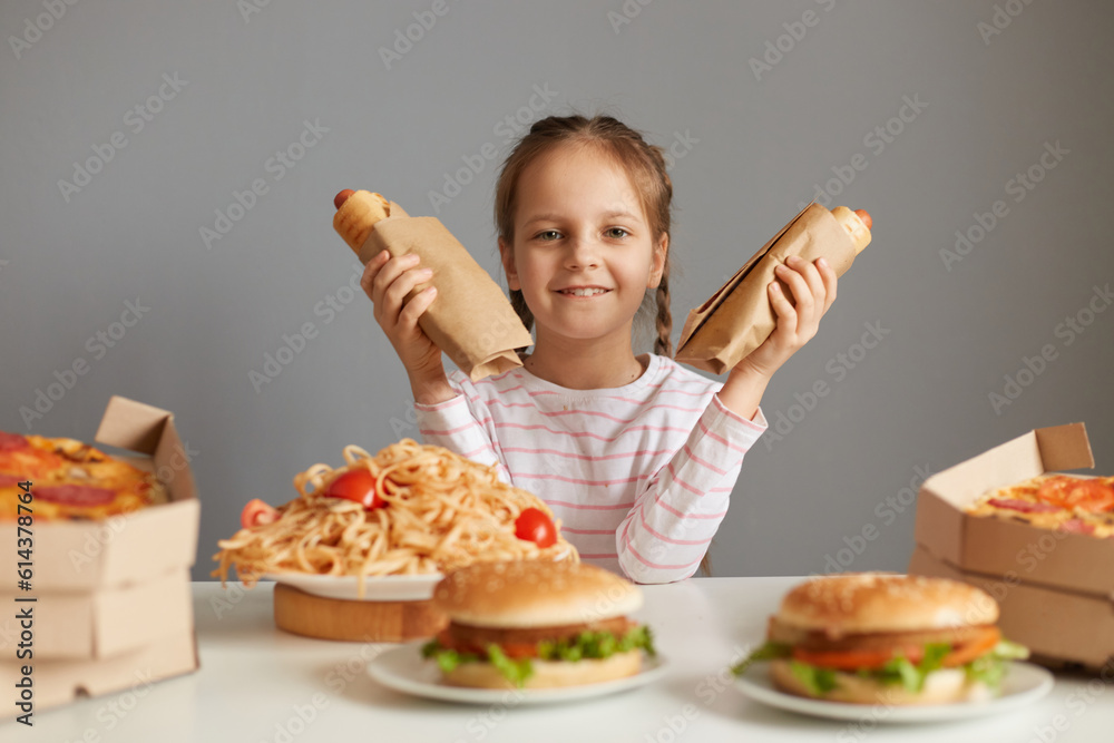 Satisfied delighted little girl with braids sitting at table with fast food isolated over gray background holding two hot dogs looking at cmaera with happy expression.