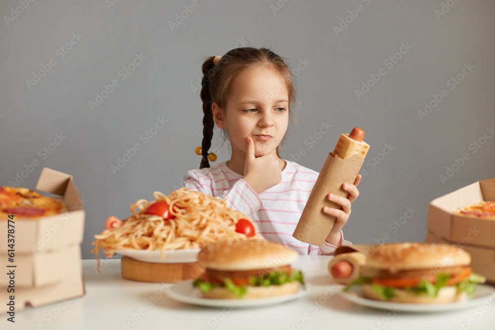 Pensive little girl with braids sitting at table with fast food isolated over gray background holding hot dog thinking eat or not holding chin looking at unhealthy meal. Pensive little girl with braids sitting at table with fast food isolated over gray background holding hot dog thinking eat or not holding chin looking at unhealthy meal.