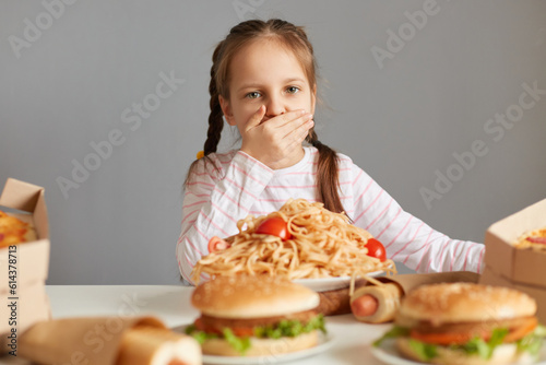 Sick ill little girl with braids sitting at table with junk food isolated over gray background feeling overating being seek feels nausea covering mouth with plam vomit.